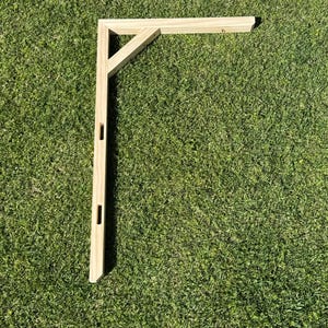 May include: A wooden bracket with a right-angle design, featuring a vertical support with rectangular cutouts and a horizontal arm. The bracket is made of light-colored wood and is set against a green grass background.