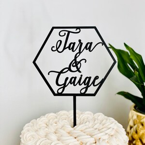 May include: Black hexagonal cake topper with the text "Tara & Gaigue" in a cursive font. The topper is on top of a white cake with a rosette design.