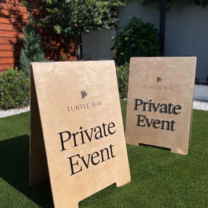 May include: Two light-colored wooden A-frame signs standing on green turf. Each sign features a small turtle icon above the text "TURTLE BAY" and the words "Private Event" in large, dark, bold lettering printed on the front surface.