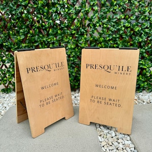 May include: Two wooden A-frame signs with black lettering. The signs read "Presqu'ile Winery" and "Welcome, Please Wait to Be Seated."