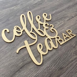 May include: Wooden sign with the words "Coffee & Tea Bar" in a cursive font. The sign is painted gold and has a rustic look.
