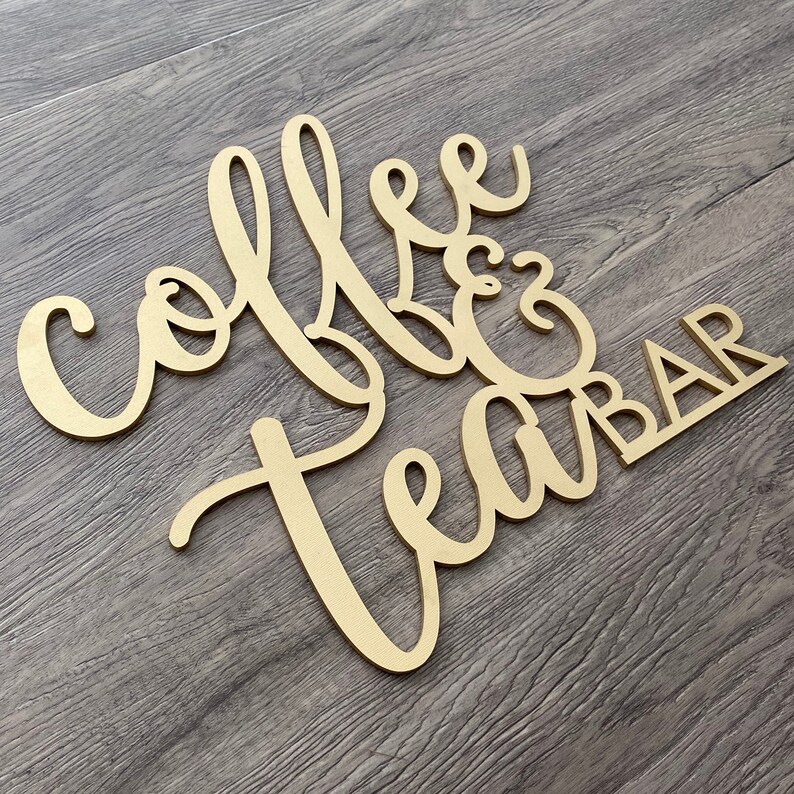 May include: Wooden sign with the words "Coffee & Tea Bar" in a cursive font. The sign is painted gold and has a rustic look.
