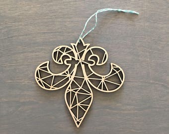 May include: A wooden fleur-de-lis ornament with a geometric design. The ornament is light brown with a light blue and white string for hanging. The fleur-de-lis is a symbol of royalty and is often used in heraldry and design.