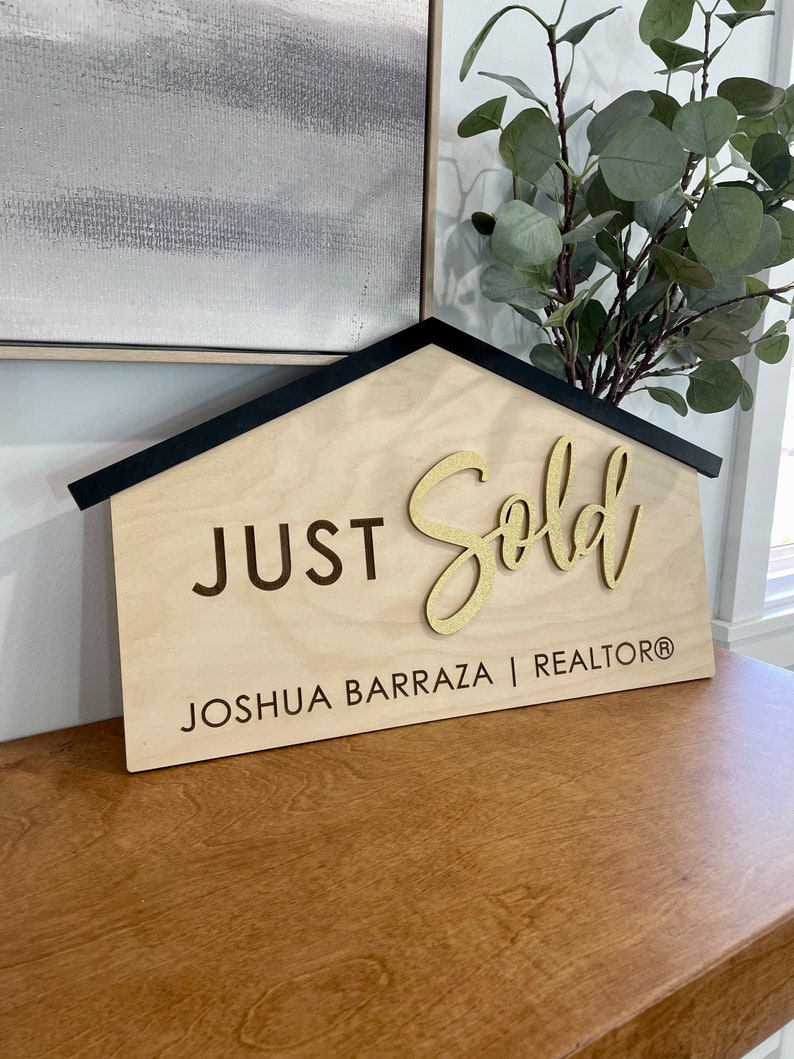 May include: A wooden house-shaped sign with black trim reads "JUST Sold" in gold glitter lettering. Below the text is "JOSHUA BARRAZA | REALTOR&reg;" in black lettering.