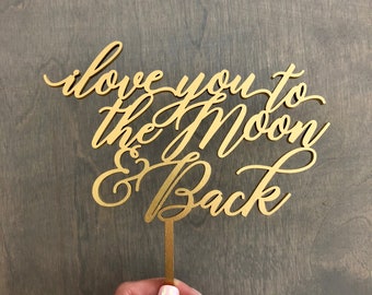 May include: Gold-colored wooden cake topper with the text "i love you to the moon & back" in a cursive font.