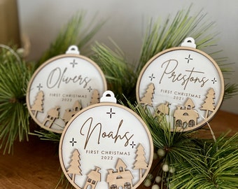 May include: Three wooden Christmas ornaments with a white background and a natural wood frame. Each ornament has a name, "First Christmas" and a year. The ornaments feature a cut-out design of houses and trees. The ornaments are hanging on a green pine branch.