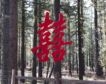 May include: A red Chinese double happiness symbol hanging from a red fabric canopy in a forest setting. The symbol is a traditional Chinese character that represents good fortune and happiness in marriage.