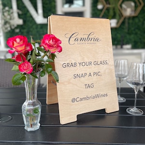 May include: A wooden sign with the text "Cambria Estate Winery" and the message "Grab your glass, snap a pic, tag @CambriaWines". The sign is on a black table with a vase of pink roses.