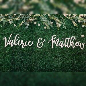 May include: A green hedge wall decorated with white flowers and greenery. The names "Valerie & Matthew" are displayed in elegant white cursive script, suitable for a wedding or event backdrop.