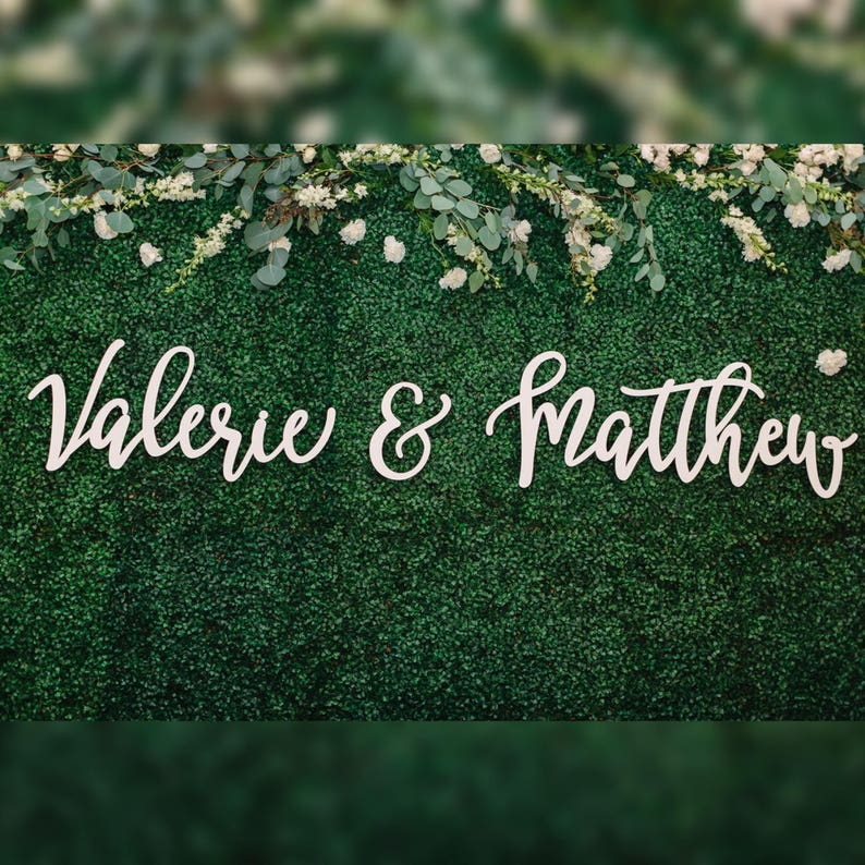 May include: A green hedge wall decorated with white flowers and greenery. The names "Valerie & Matthew" are displayed in elegant white cursive script, suitable for a wedding or event backdrop.