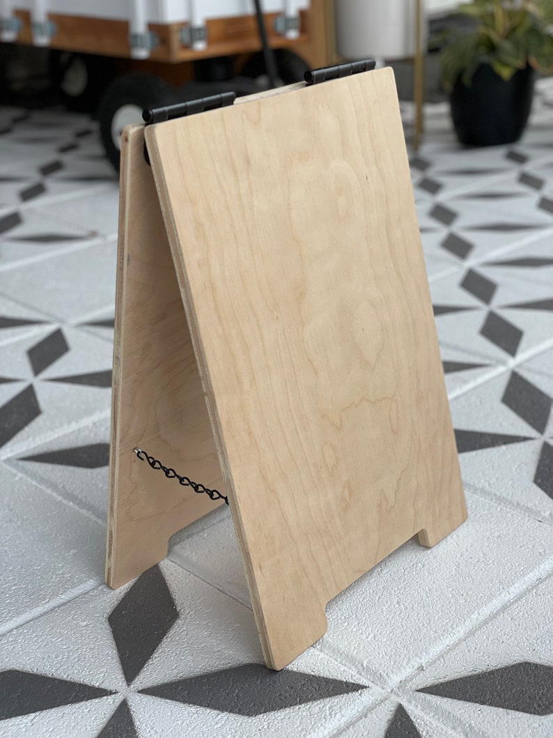 May include: A wooden A-frame sign with black hinges and a chain. The sign is made of light-colored wood and is standing on a tiled floor.