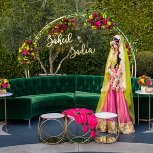 May include: An outdoor wedding scene with a green velvet sofa, gold side tables, and a floral arch. A person in a pink and gold outfit stands near the sofa. The arch has the names "Sahil + Sadia" in gold script.