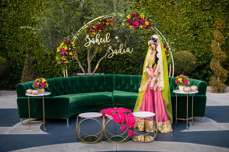 May include: An outdoor wedding scene with a green velvet sofa, gold side tables, and a floral arch. A person in a pink and gold outfit stands near the sofa. The arch has the names "Sahil + Sadia" in gold script.