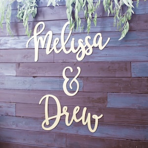 May include: Wooden sign with the names "Melissa & Drew" in elegant cursive script, mounted on a dark brown wooden wall. Eucalyptus greenery drapes across the top, adding a natural touch.