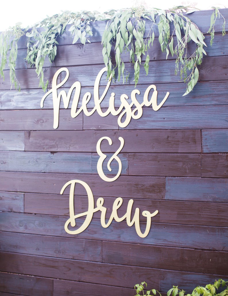 May include: Wooden sign with the names "Melissa & Drew" in elegant cursive script, mounted on a dark brown wooden wall. Eucalyptus greenery drapes across the top, adding a natural touch.