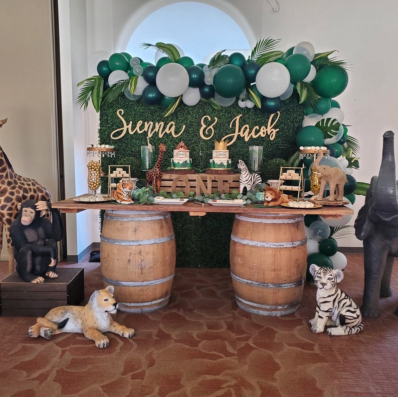 May include: A jungle-themed party setup with a wooden table supported by barrels. The backdrop features a green wall with balloons, greenery, and the names "Sienna & Jacob." Decorative animal figurines and cakes are displayed.