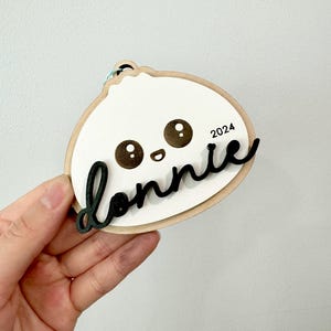 May include: A wooden ornament shaped like a cartoon dumpling with a smiling face and the name "Donnie" written in black cursive lettering. The year "2024" is printed in black on the ornament.