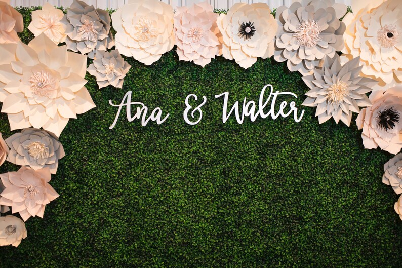 May include: A decorative backdrop featuring a green textured surface, adorned with large paper flowers in shades of cream, pink, and gray. The names "Ana & Walter" are displayed in white script across the center.