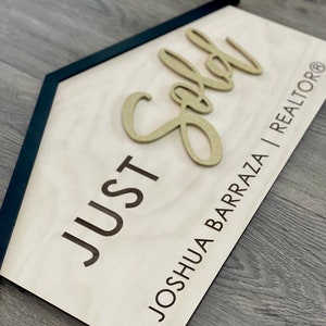 May include: A wooden sign with a black frame in the shape of a house. The sign says "JUST SOLD" in black lettering and "JOSHUA BARRAZA | REALTOR&reg;" in smaller black lettering. The word "SOLD" is in gold glitter lettering.