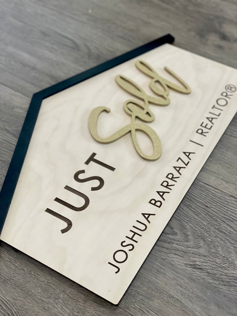 May include: A wooden sign with a black frame in the shape of a house. The sign says "JUST SOLD" in black lettering and "JOSHUA BARRAZA | REALTOR&reg;" in smaller black lettering. The word "SOLD" is in gold glitter lettering.