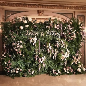May include: A lush green floral wall with a variety of flowers in shades of pink, white, and purple. The names "Lilian & Anthony" are displayed in elegant cursive script, creating a romantic and elegant backdrop.