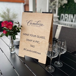 May include: A wooden sign with the text "Cambria Estate Winery" and the message "Grab your glass, snap a pic, tag @CambriaWines". Two wine glasses are on a black table in front of the sign.