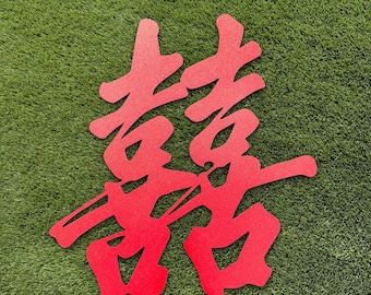 May include: A bright red double happiness symbol cut from a flat material. The Chinese character features a symmetrical design with bold, connected strokes, creating a singular decorative piece with a textured surface finish throughout the entire shape.