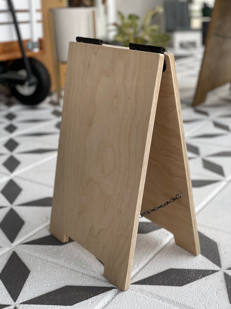May include: A wooden A-frame sign with a chain attached to the back. The sign is made of light-colored wood and has a natural finish. The sign is standing on a black and white tiled floor.