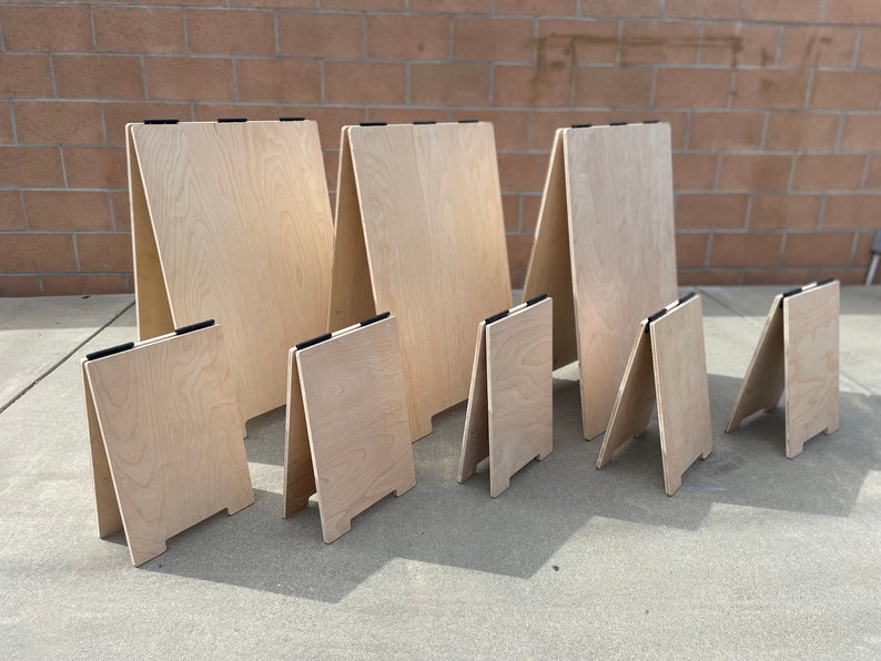May include: A set of seven wooden A-frame signs of varying sizes. The signs are made of light-colored wood and have black clips at the top.