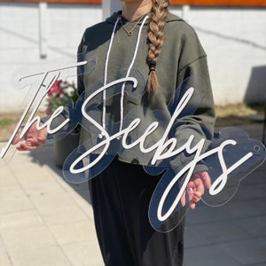 May include: A clear acrylic sign with the text "The Seeby's" in white script lettering. The sign is being held by a person wearing a green hoodie.