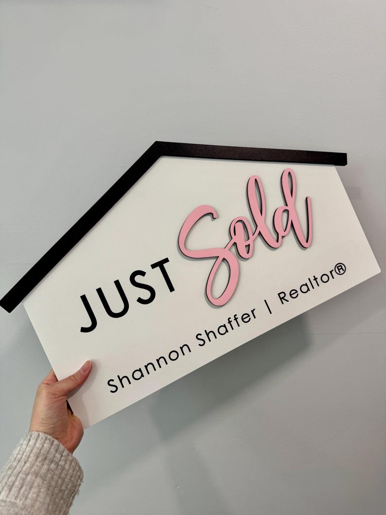 May include: A white house-shaped sign with black trim reads "JUST SOLD" in pink lettering. Below the text is "Shannon Shaffer | Realtor&reg;" in black lettering.