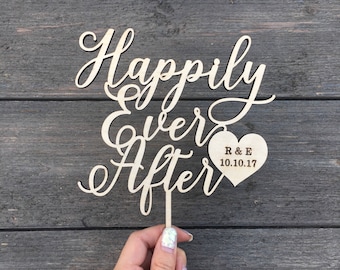 May include: Wooden cake topper with the words "Happily Ever After" in cursive script. A small heart-shaped tag is attached to the topper with the text "R & E 10.10.17" engraved on it.