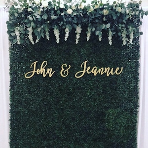May include: A dark green boxwood backdrop with the names "John & Jeannie" in gold script. The top of the backdrop is decorated with white flowers, green leaves, and white hanging floral accents. The backdrop is likely for a wedding or event.