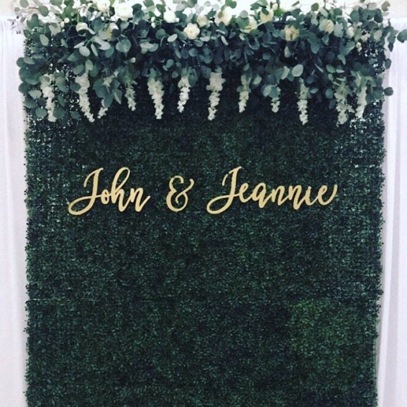 May include: A dark green boxwood backdrop with the names "John & Jeannie" in gold script. The top of the backdrop is decorated with white flowers, green leaves, and white hanging floral accents. The backdrop is likely for a wedding or event.