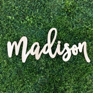 May include: A wooden sign with the name "Madison" written in cursive script. The sign is made of natural wood and has a light brown color.