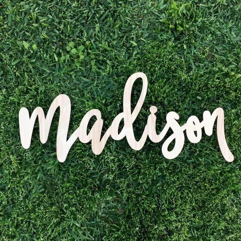 May include: A wooden sign with the name "Madison" written in cursive script. The sign is made of natural wood and has a light brown color.