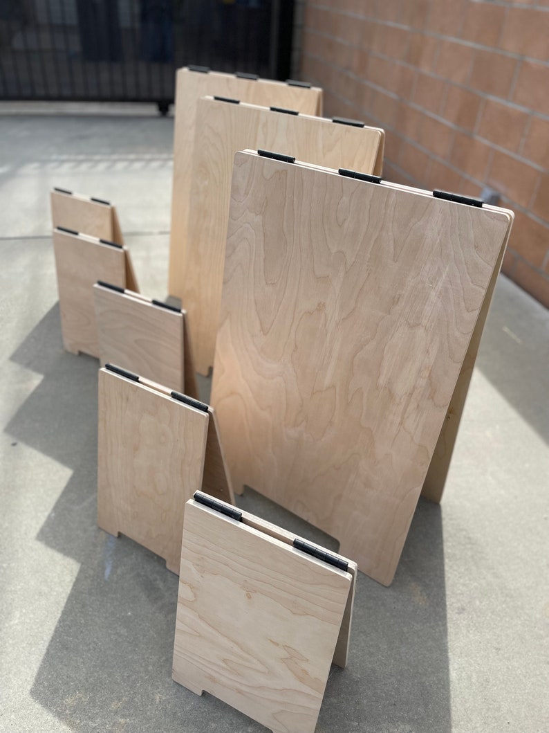 May include: A set of six wooden A-frame signs in various sizes. The signs are made of light-colored wood with a natural grain pattern. The signs are all standing upright on a concrete surface.