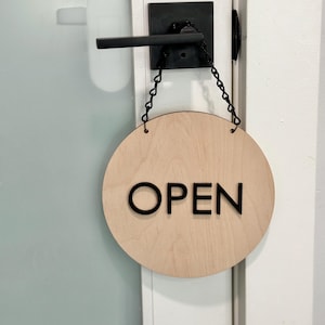 May include: A round wooden sign with the word "OPEN" in black lettering hangs from a black chain on a white door.