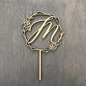 May include: Wooden cake topper with a geometric frame and the letter "M" in a cursive font. The frame is decorated with floral details.