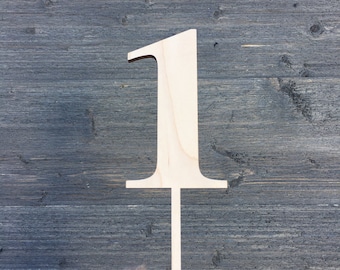 May include: A wooden number one cutout, painted white, with a stick attached to the bottom. The number is held in a hand wearing a brown knitted glove.