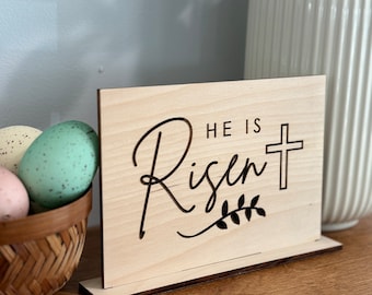 May include: Wooden sign with the text "He is Risen" and a cross with a leafy branch underneath. The sign is standing on a wooden base.