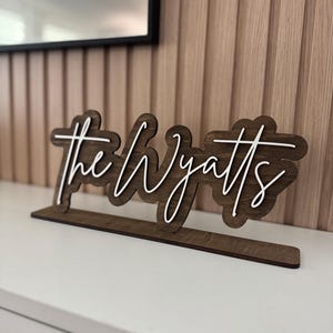 May include: Wooden sign with the text "the Wyatts" in a cursive font. The sign is brown with white lettering and sits on a white surface. The sign is a decorative item.