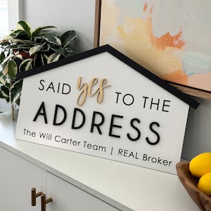 May include: A white house-shaped sign with black trim reads "Said yes to the address The Will Carter Team | REAL Broker". The sign has a gold "yes" written in cursive.