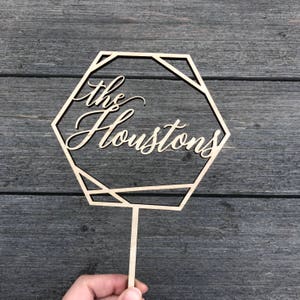 May include: Wooden cake topper with a geometric hexagon design. The topper features the text "the Houstons" in a cursive font.
