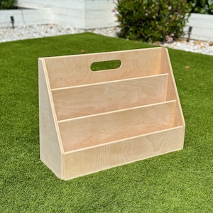May include: A wooden book organizer with three shelves and a handle on the top. The organizer is made of light wood and is sitting on a green artificial grass surface.
