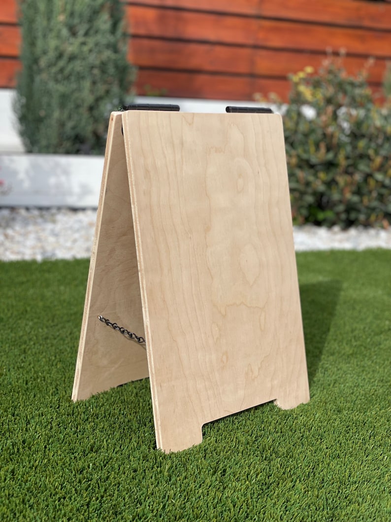 May include: A blank wooden A-frame sign with black metal hinges. The sign is made of light-colored wood and is standing on a green artificial grass surface.