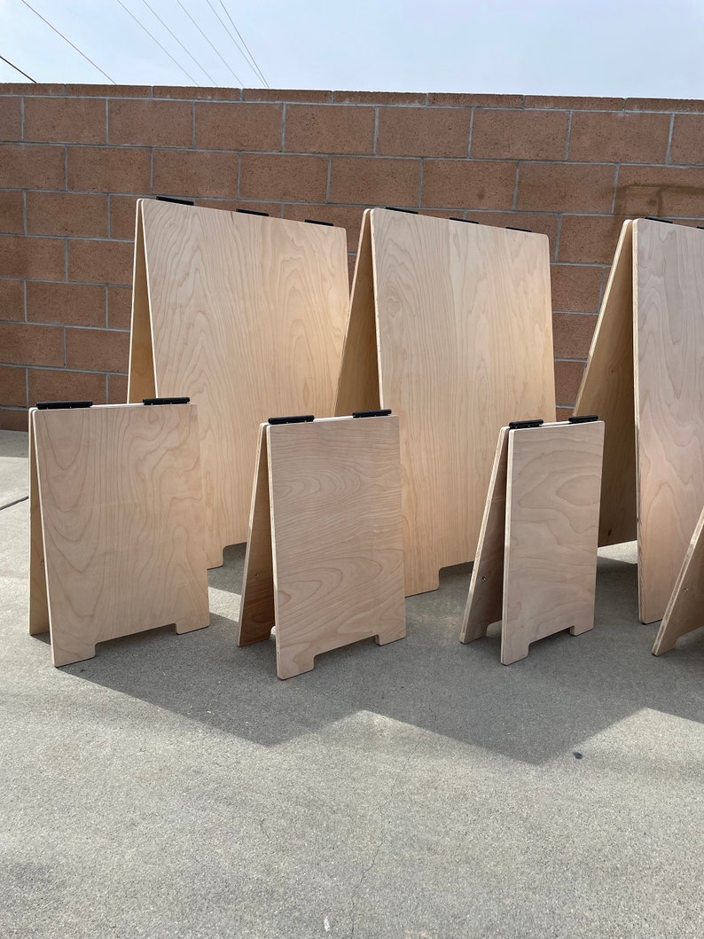 May include: Five wooden A-frame signs of varying sizes, all made of light-colored wood with black hinges. The signs are standing on a gray concrete surface.