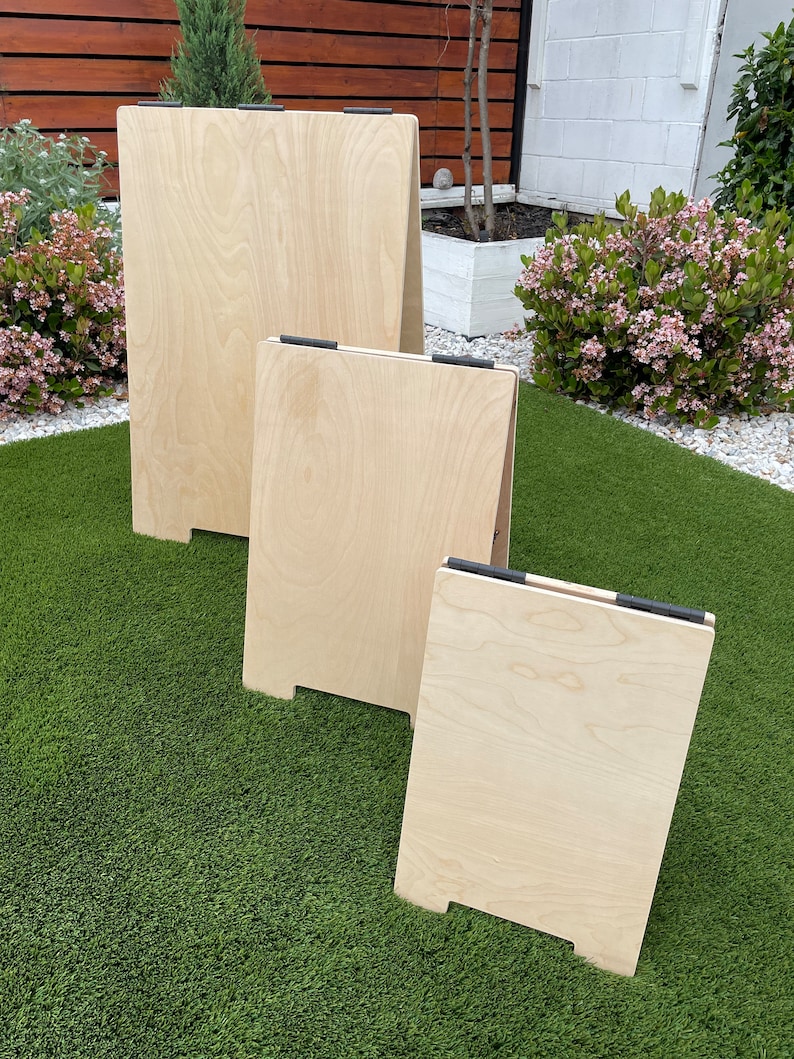 May include: Three wooden A-frame signs of varying sizes, all made of light-colored wood with black hinges. The signs are standing on a green artificial grass surface.