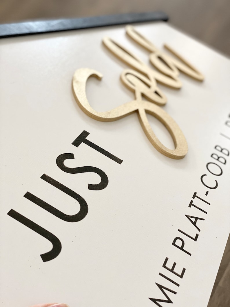 May include: A white wooden sign with black text that reads "JUST" and a gold wooden cutout of the word "Gold". The sign is framed with a black frame and has the text "JAMIE PLATT-COBB" in black text at the bottom.
