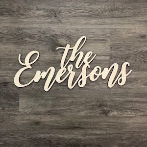 May include: The Emersons family name in cursive script, cut out of wood and painted white, against a dark wood background.